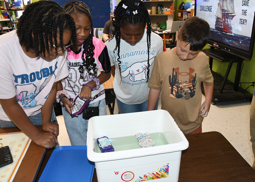 Four children with various hairstyles, one holding a cell phone, look at a small boat in a bin of water.