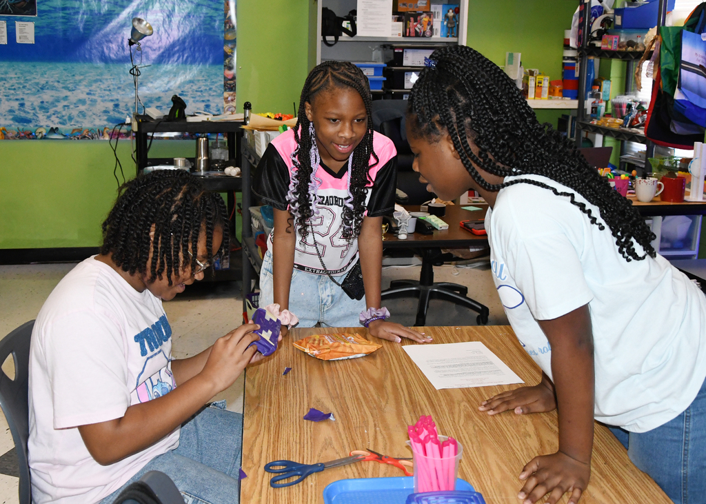 Three children collaborate around a desk; one is seated and holding an object while the others watch. Background includes shelves and a mural.