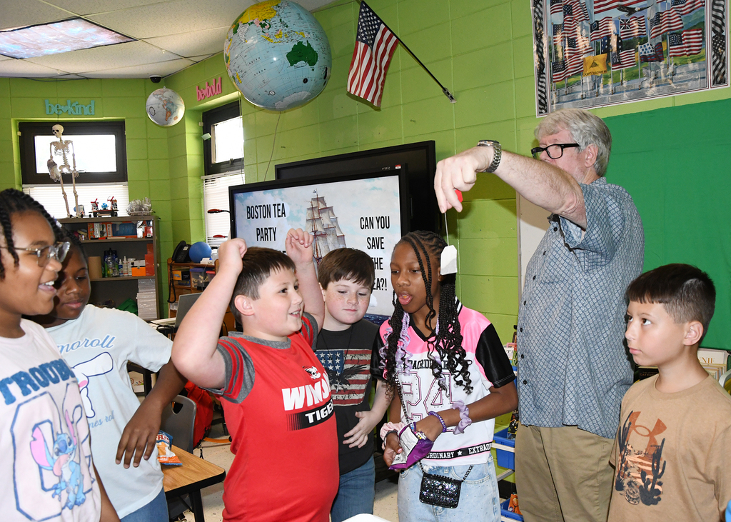 A group of children and an adult male teacher in a classroom with green walls.