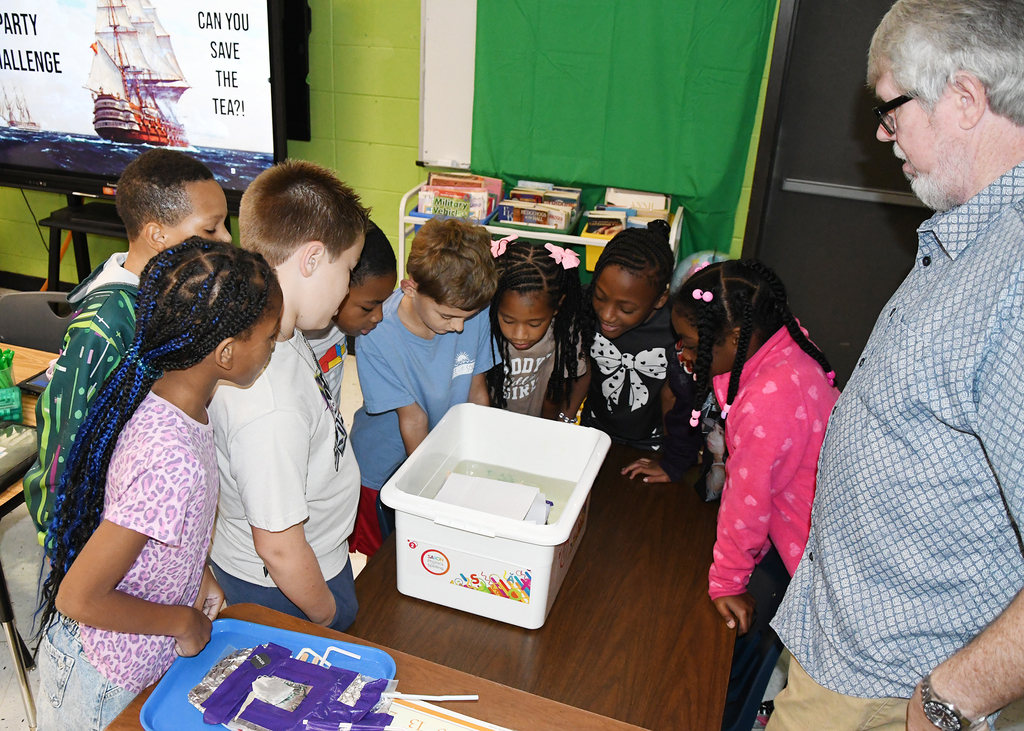 A group of children are gathered around a table with a box on it. An adult man is also present.