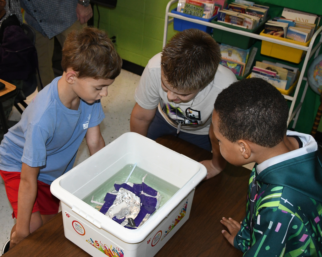 Three children focus on a container with water and floating objects on a table in a classroom.