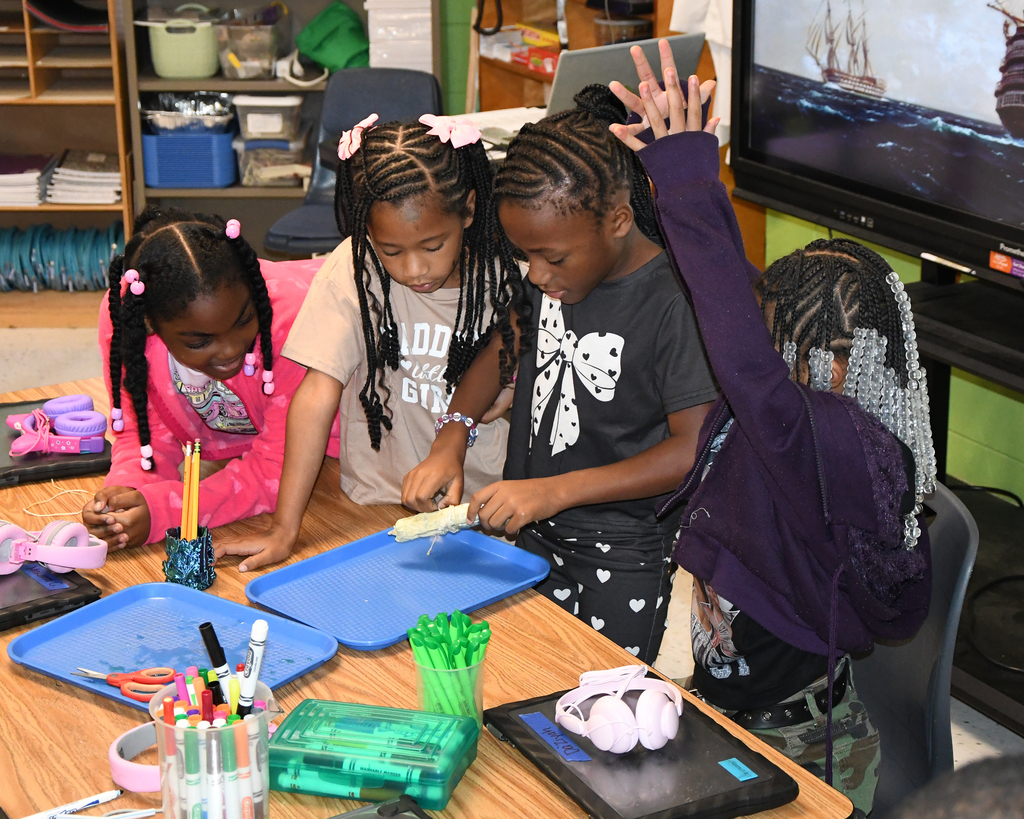 Four girls seated at a table in a classroom. One is raising her hand. Behind them, a monitor shows a picture of a ship.