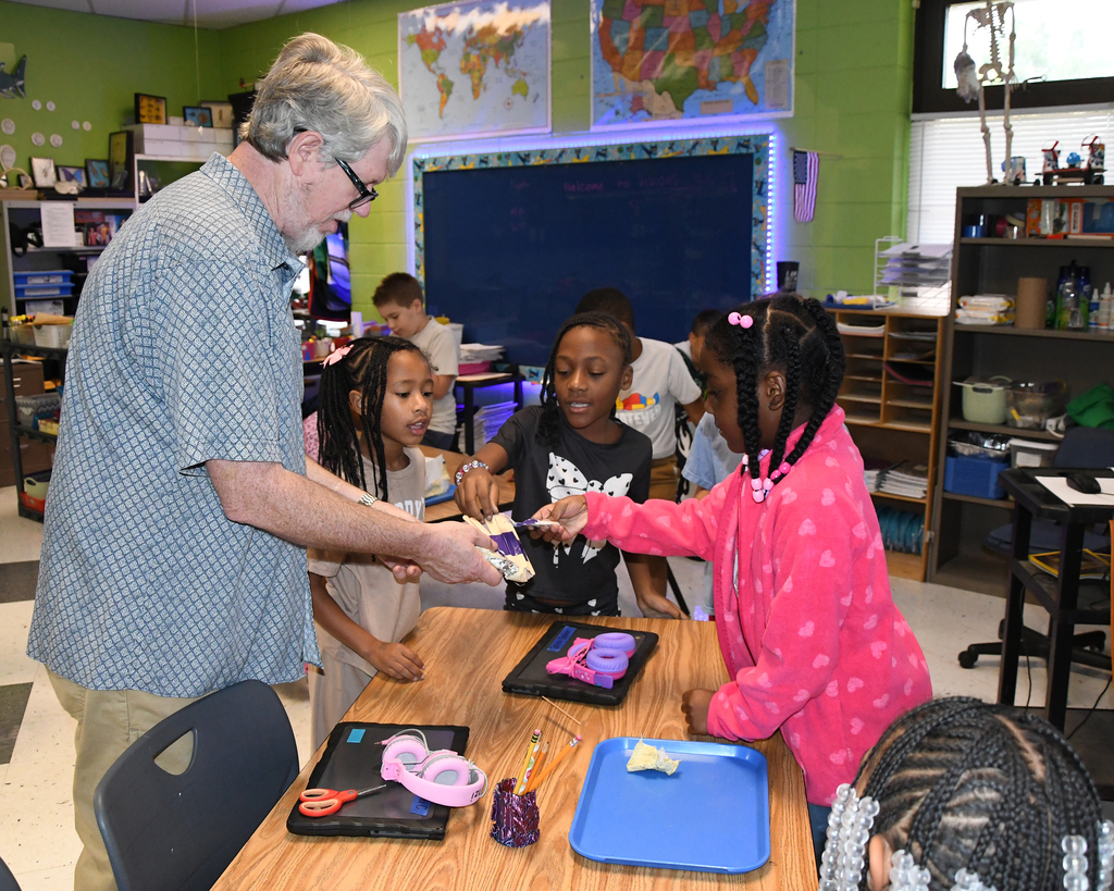An adult man gives a girl a cookie from a tray while others watch in a classroom.