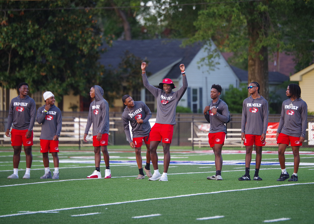 Nine men wearing gray shirts and red shorts stand on a field, with one raising his hands.