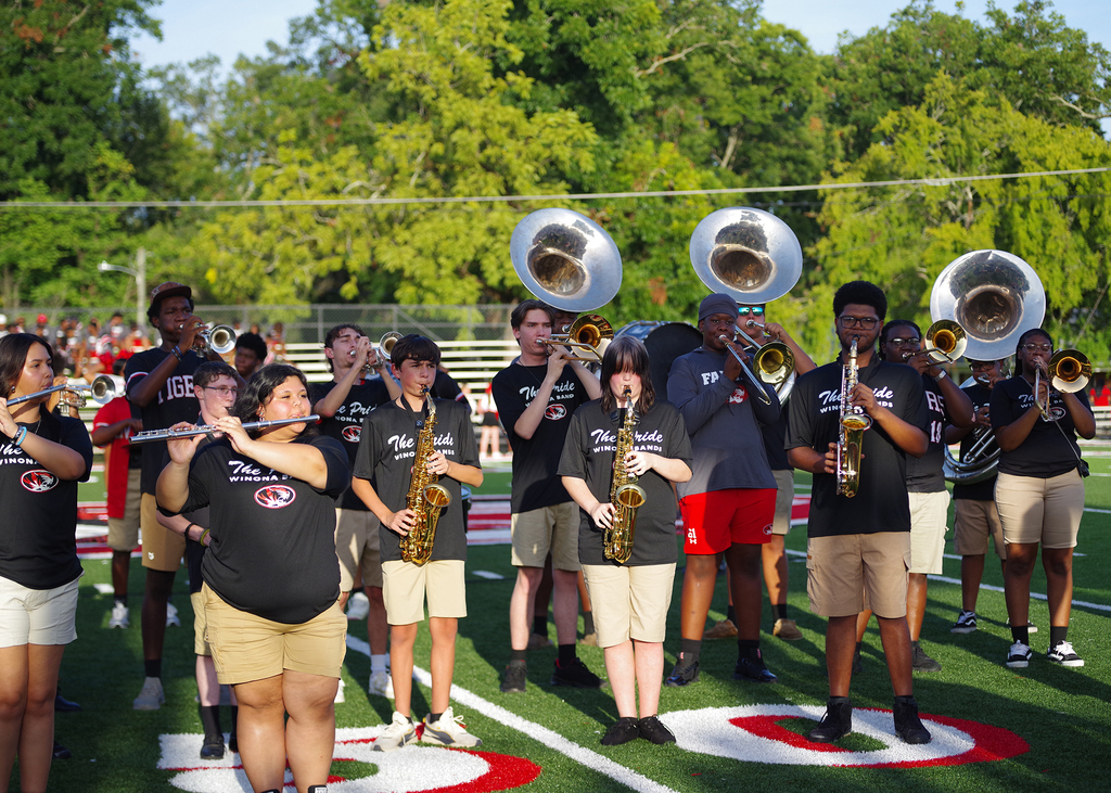 A school band practices on a field with green grass and trees. They play various instruments, including saxophones and tubas.