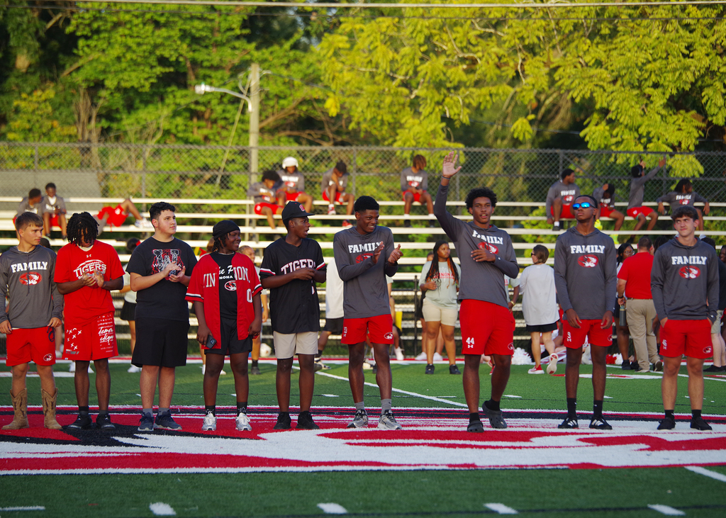 A group of students in sports uniforms stand on a football field with a red and white logo.