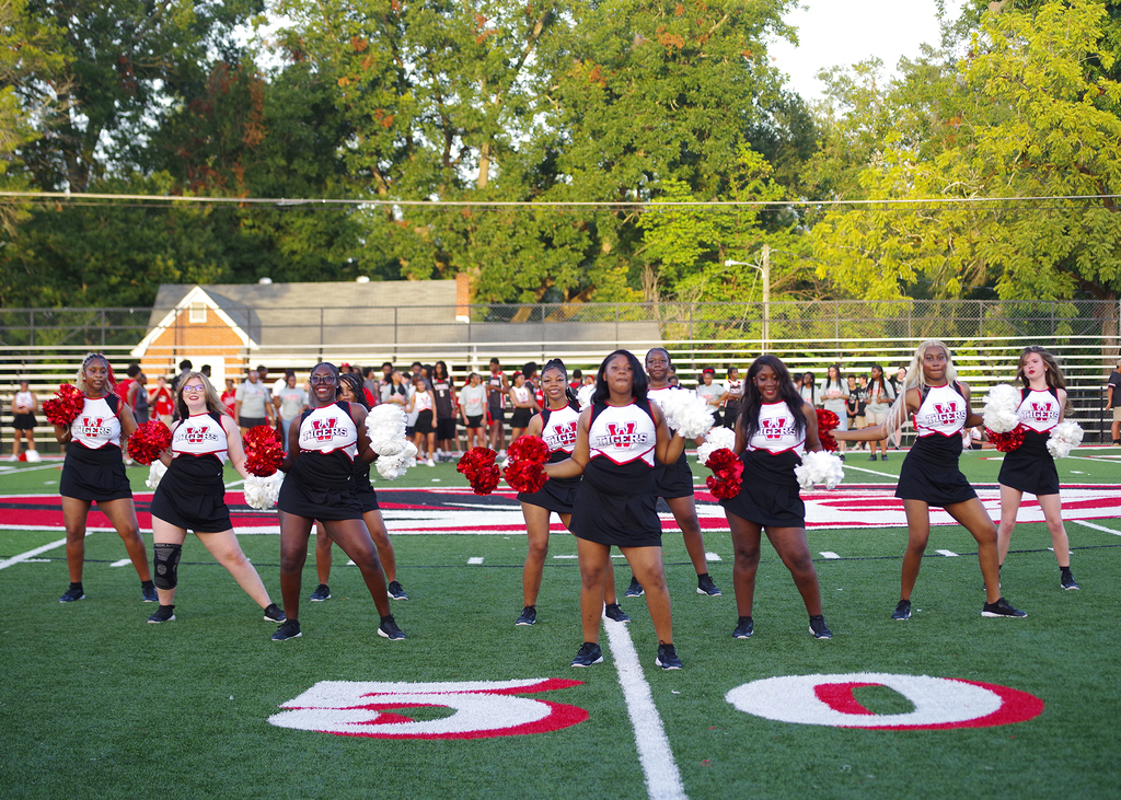 Cheerleaders in black and red uniforms perform on a field, with red and white pom-poms. Trees and a house in the background.