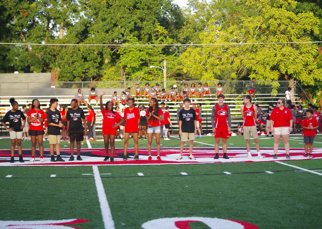 People in red shirts and shorts stand on a football field, facing the bleachers with trees in the background.