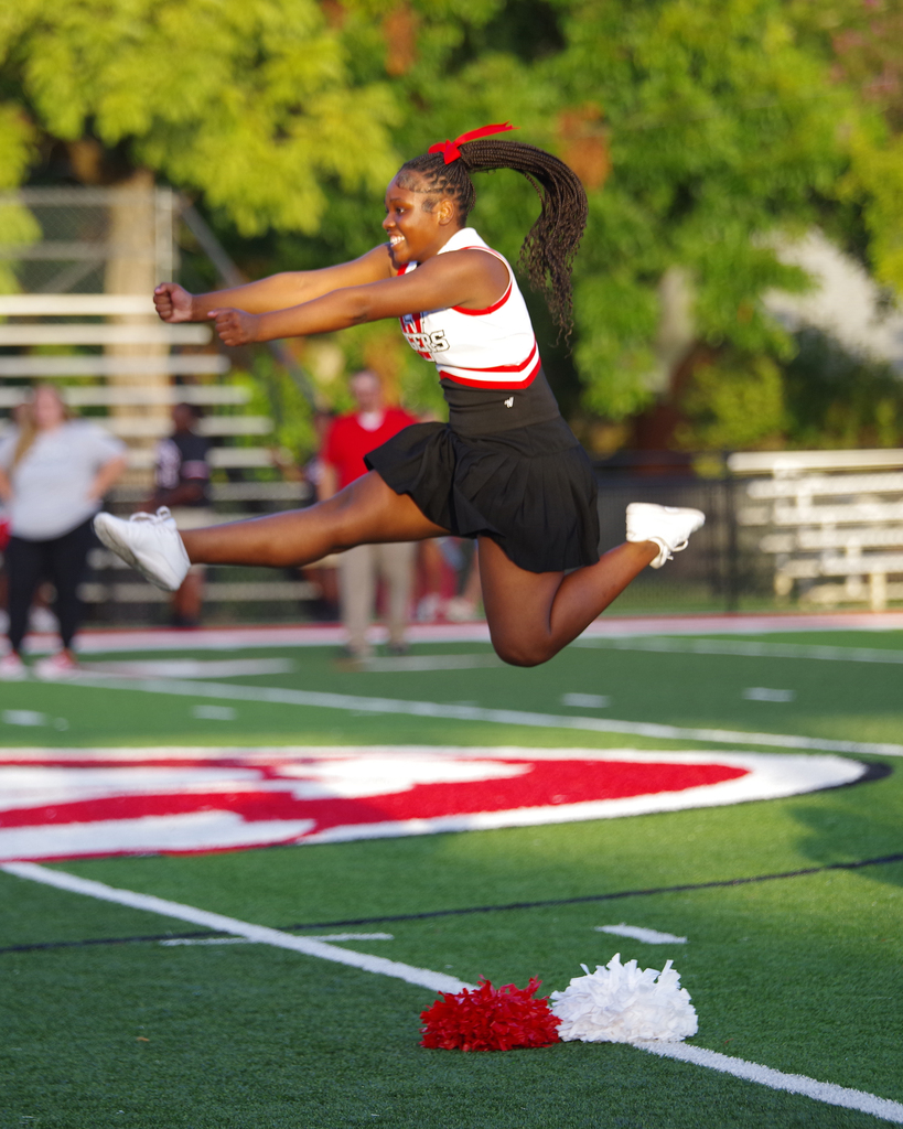 Person jumping in air with legs spread, holding pom poms. Behind, field with red and white logo.