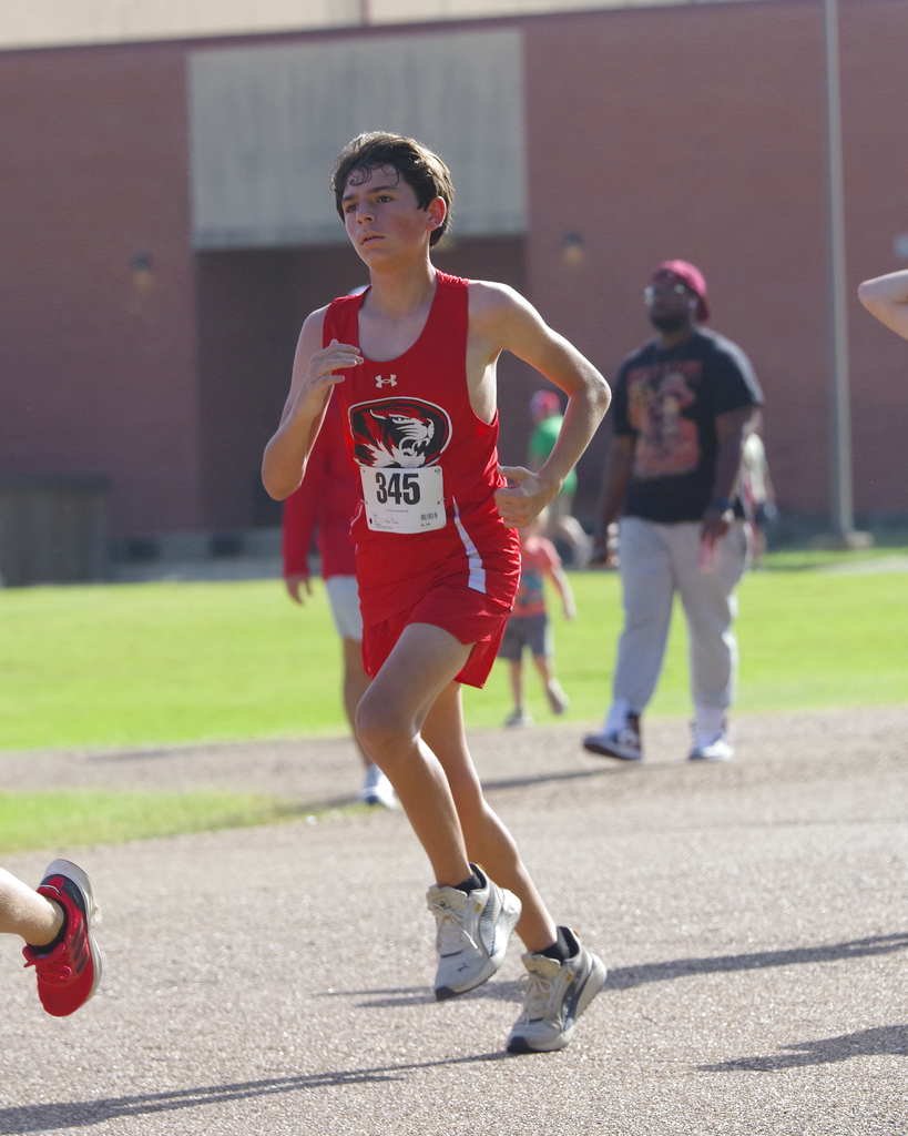 A runner with a red number 345 on his shirt leads in a race on a paved track, with a grassy area behind.