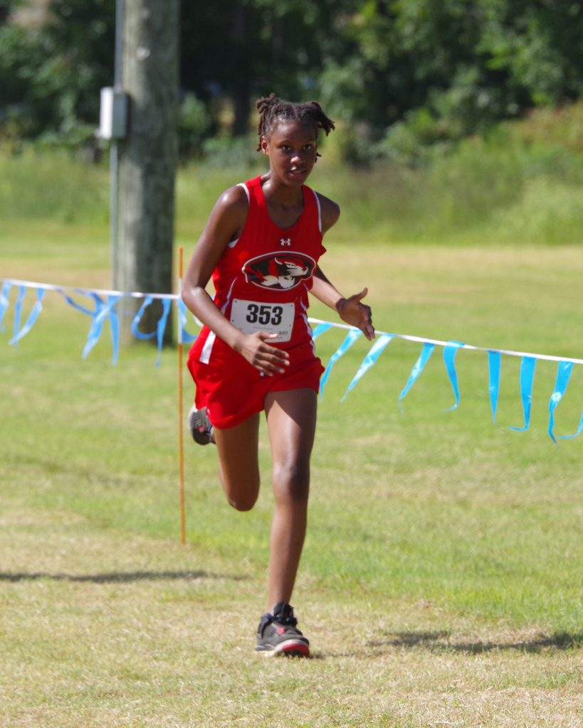 A female athlete runs in a cross-country race with number 353 on her shirt. She is surrounded by a grassy field and a blue ribbon barrier.
