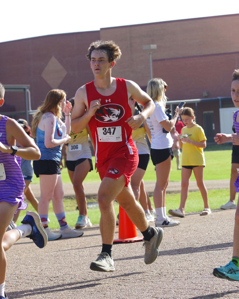 A boy wearing a red shirt and number 347 runs in a race. Spectators in various shirts watch behind.