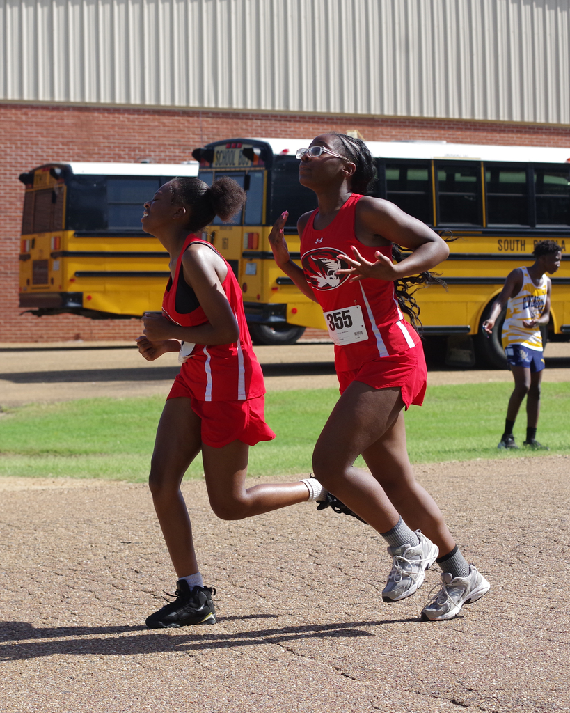 Two women in red uniforms run side by side on a track. A yellow school bus and a man stand on grass in the background.