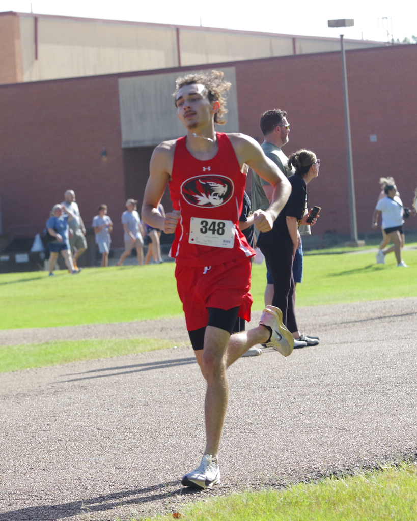 A man wearing a red shirt and black shorts runs on a track with a number 348. People walk behind him on a grassy field.