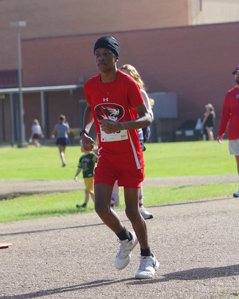 A man in red athletic clothing runs on a track with a building in the background.