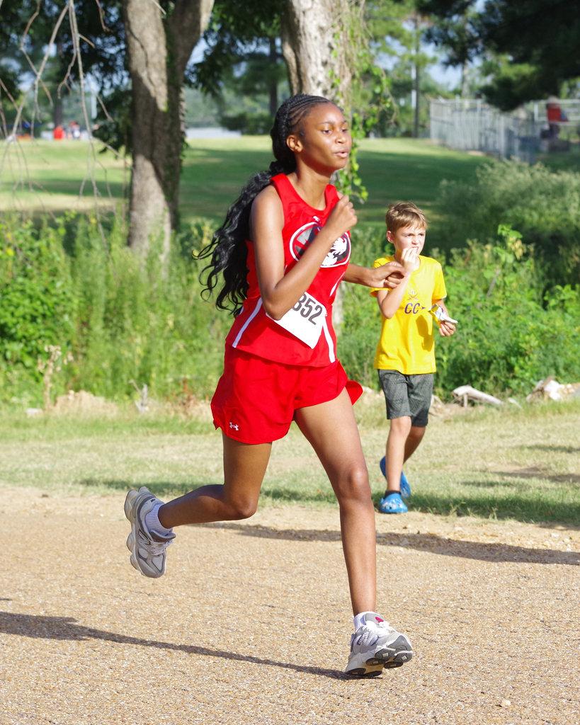 A girl with a race number running with a boy in a park with green bushes.