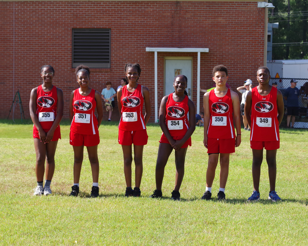 Five individuals in red track uniforms with numbers stand on a grassy field. Behind them is a brick building with an entrance.