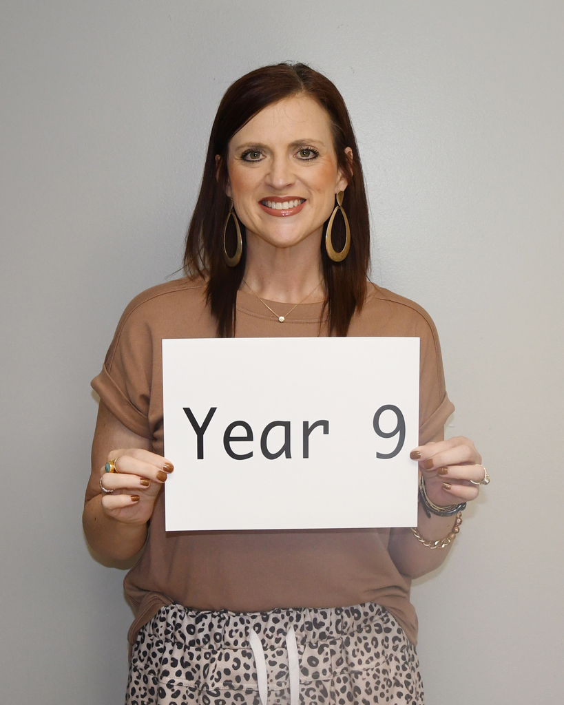 A woman in a brown top and leopard print skirt smiles while holding a white sign that says "Year 9".