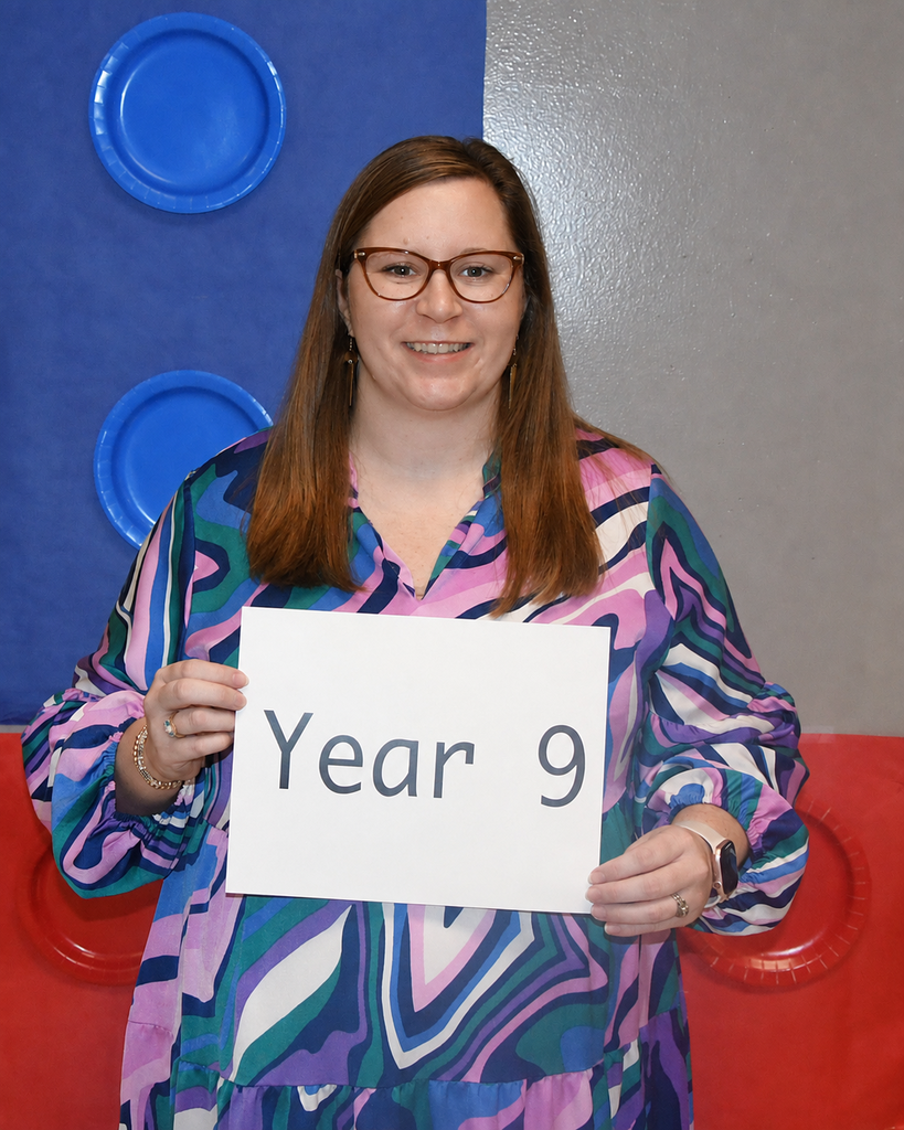 Woman holding a sign reading "Year 9" while smiling. She wears glasses and a patterned blouse. Behind her is a colorful backdrop with blue circles.