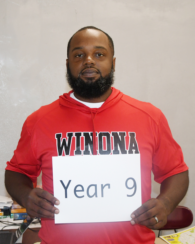 A man in a red shirt with "WINONA" stands holding a sign that says "Year 9" in front of a white background.