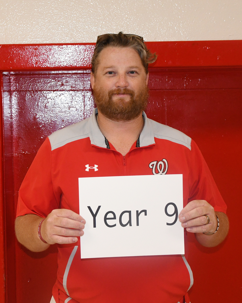 A man in red holds a white sign that reads "Year 9" in front of a red locker door.