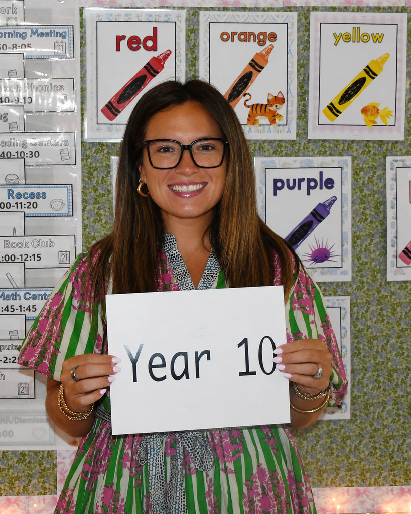 A woman wearing glasses holds a white sign reading "Year 10" in front of a classroom with colorful posters.