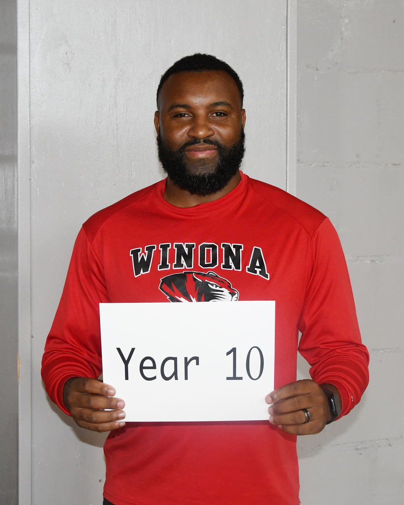 A man stands in front of a white wall, holding a white sign that reads "Year 10."