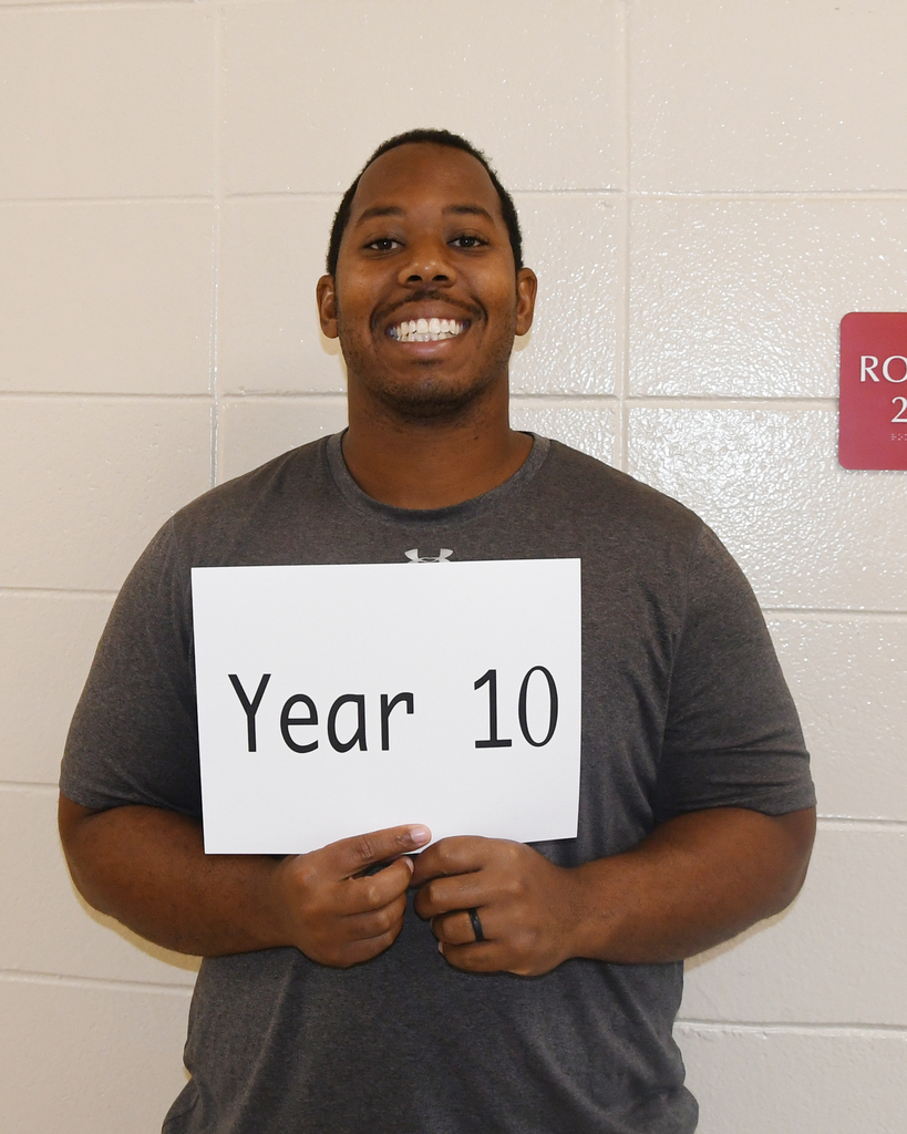 A man stands in front of a wall, holding a sign that reads "Year 10". He wears a gray T-shirt and smiles.