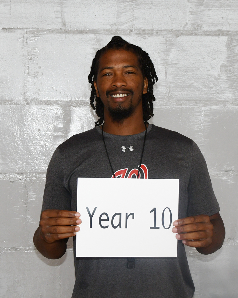 A man in a gray shirt holds a white sign with black text that reads "Year 10" against a light-colored brick wall.