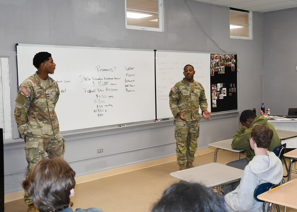 Two uniformed military personnel stand before a whiteboard. Students in a classroom setting sit at desks, listening.