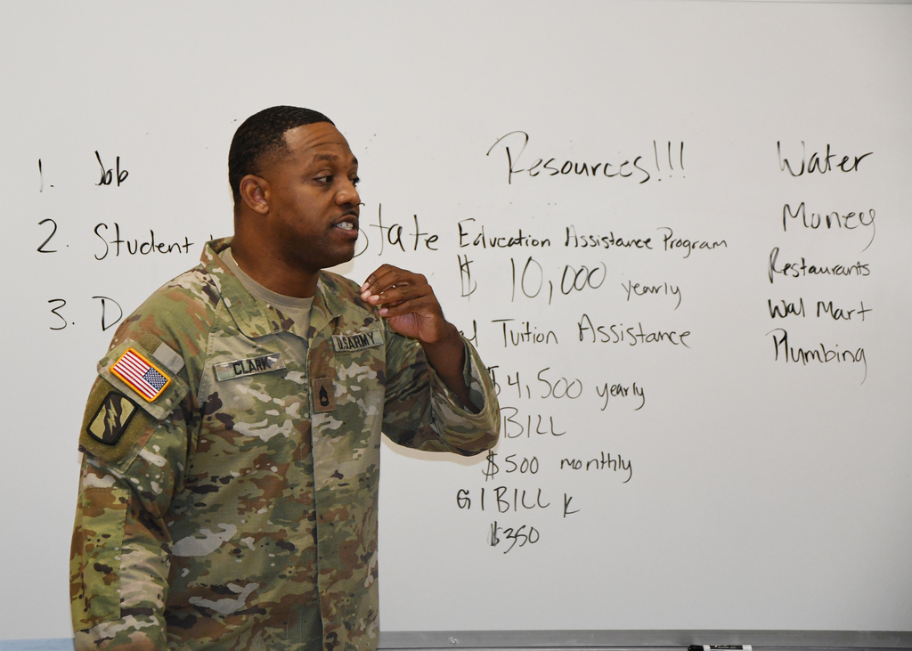 Man in uniform stands before a whiteboard with a list of financial resources and amounts.