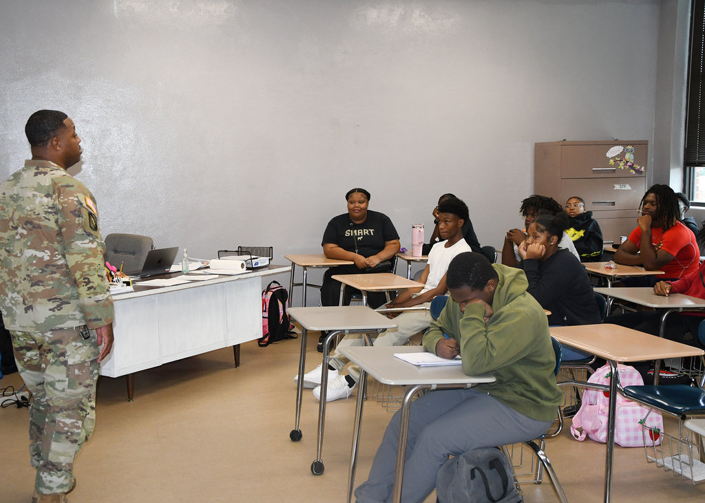 A uniformed military instructor speaks in a classroom. Students sit at desks, with some looking down. A backpack rests nearby.