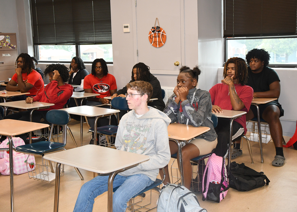 A classroom with students seated at desks, some wearing hoodies. Backpacks and bags are near the chairs.