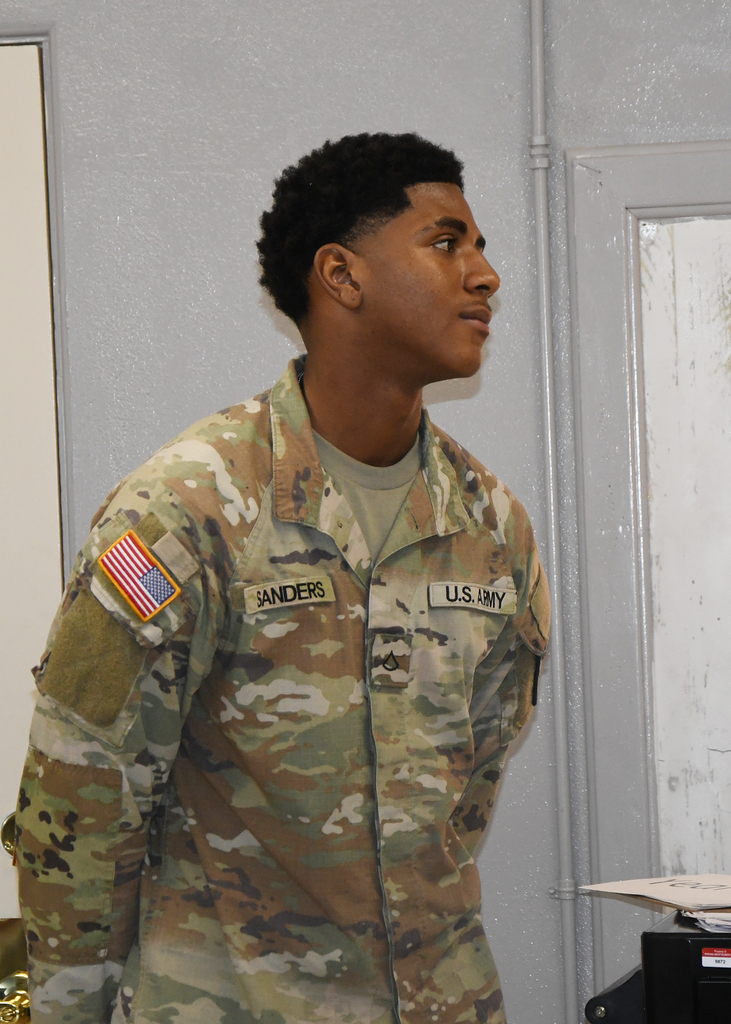 Man in U.S. Army uniform, with camouflage jacket, stands against a gray wall. An American flag patch is on his left shoulder.