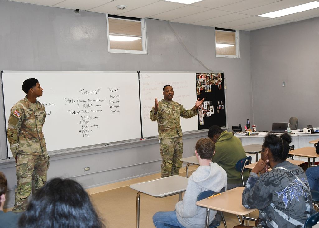 A classroom with white walls. Two military personnel stand before a whiteboard. Students sit at desks and listen.