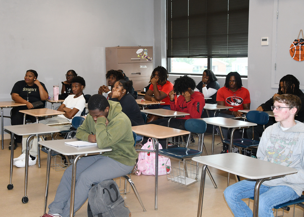 A classroom with students seated at desks. Some are facing forward; others are turned. A basketball hangs on a wall.