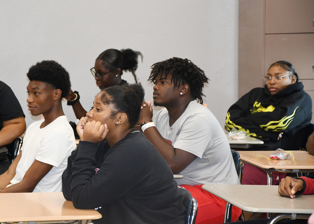 A group of students seated in a classroom, with three in front wearing black and two in white, seated at desks.