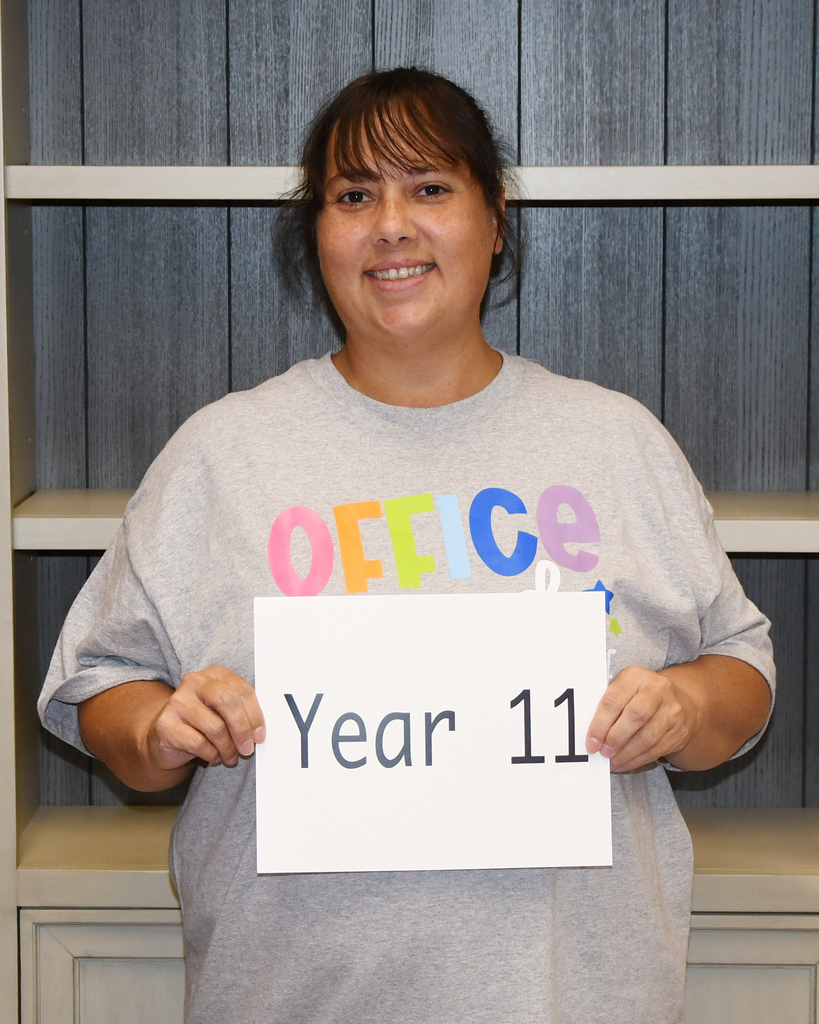 A woman holds a white sign that says "Year 11" with a smile, standing in front of a wooden cabinet.
