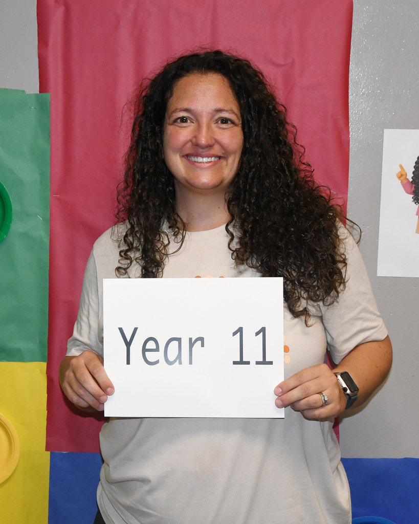A woman with curly hair holds a sign saying 'Year 11'. She is smiling and standing against a colorful backdrop.