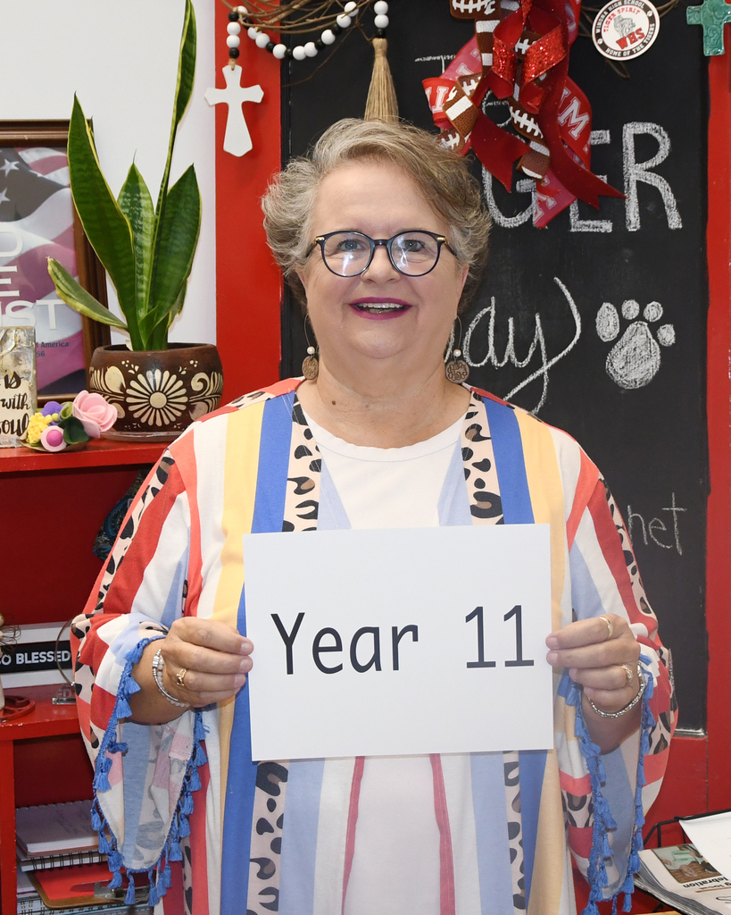 A woman with glasses stands behind a sign reading "Year 11" in a room with a blackboard and decorations.