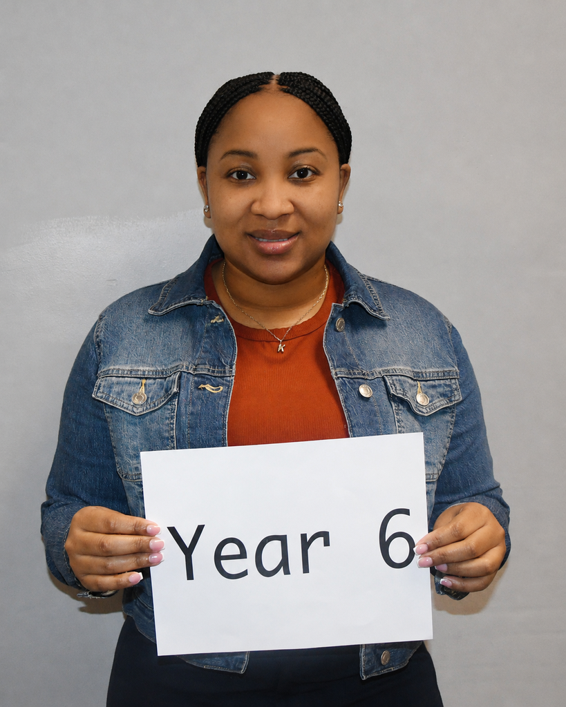 Person standing against a plain wall, wearing a denim jacket, holding a white sign reading "Year 6".