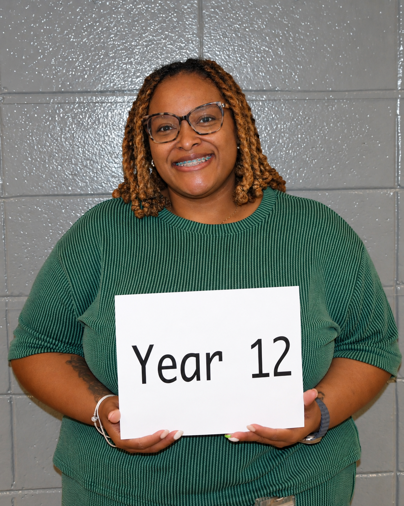Person with curly hair, wearing glasses and a green top, holding a sign that reads "Year 12".