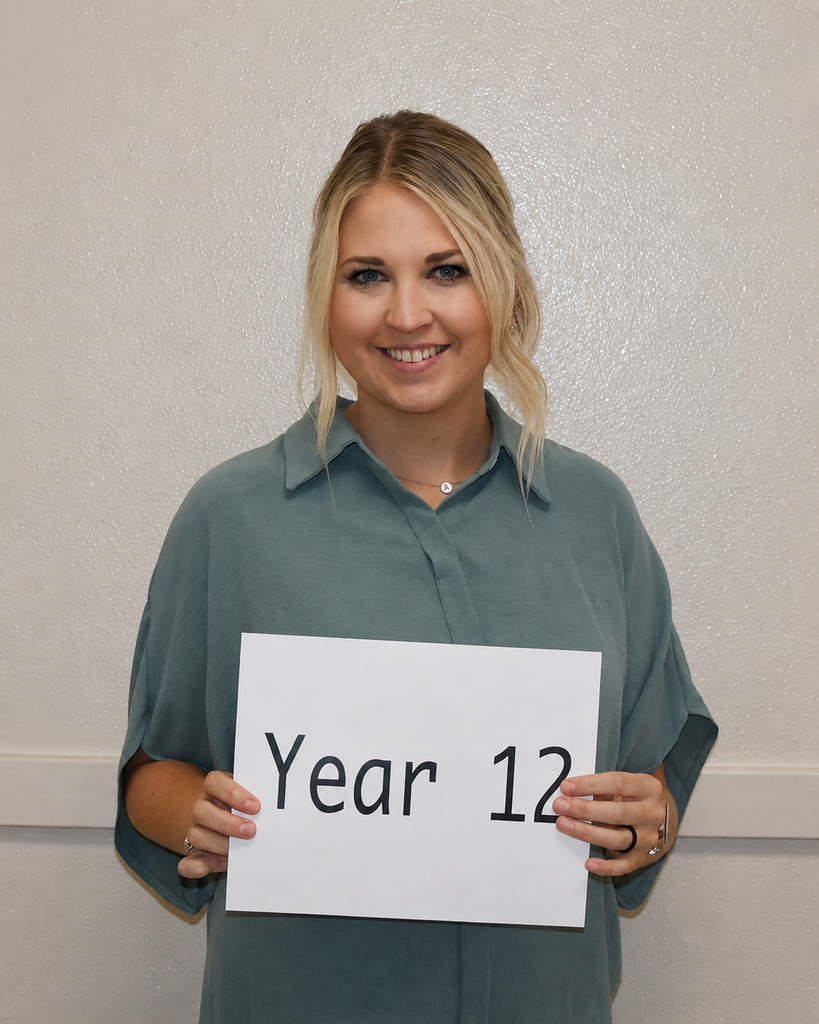 A person poses, smiling, with a white sign reading "Year 12". The person wears a green shirt, with a light-colored background.