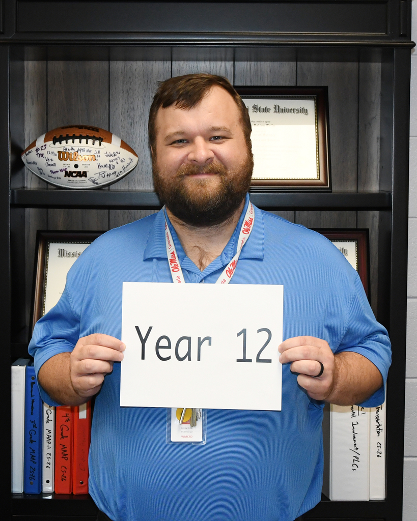 Person holds a sign that says Year 12 in front of a bookshelf. Behind them, books, a football, and framed certificates.