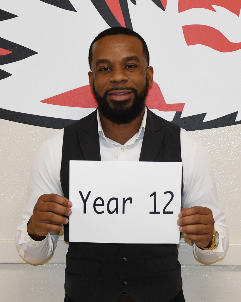 A man in a white shirt and black vest holding a sign with the text "Year 12" in front of a wall with a red and white graphic.