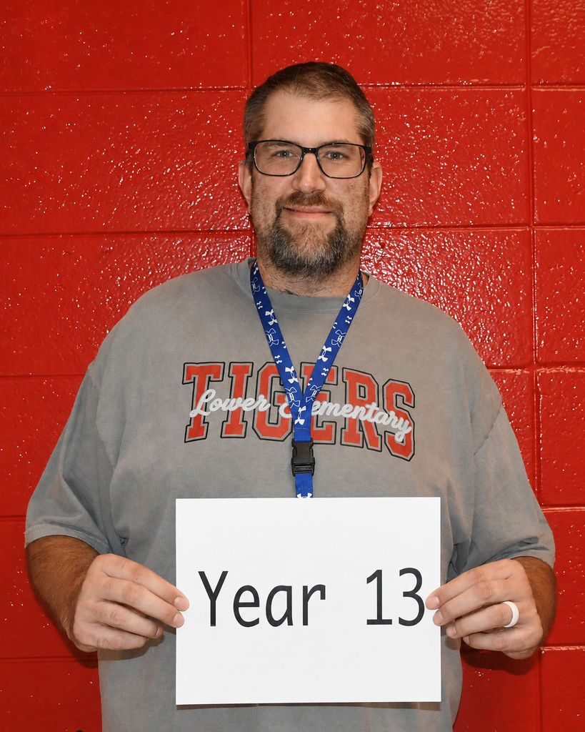 Man with a beard wearing glasses and a gray shirt with "Tigers" holds a white sign reading "Year 13".