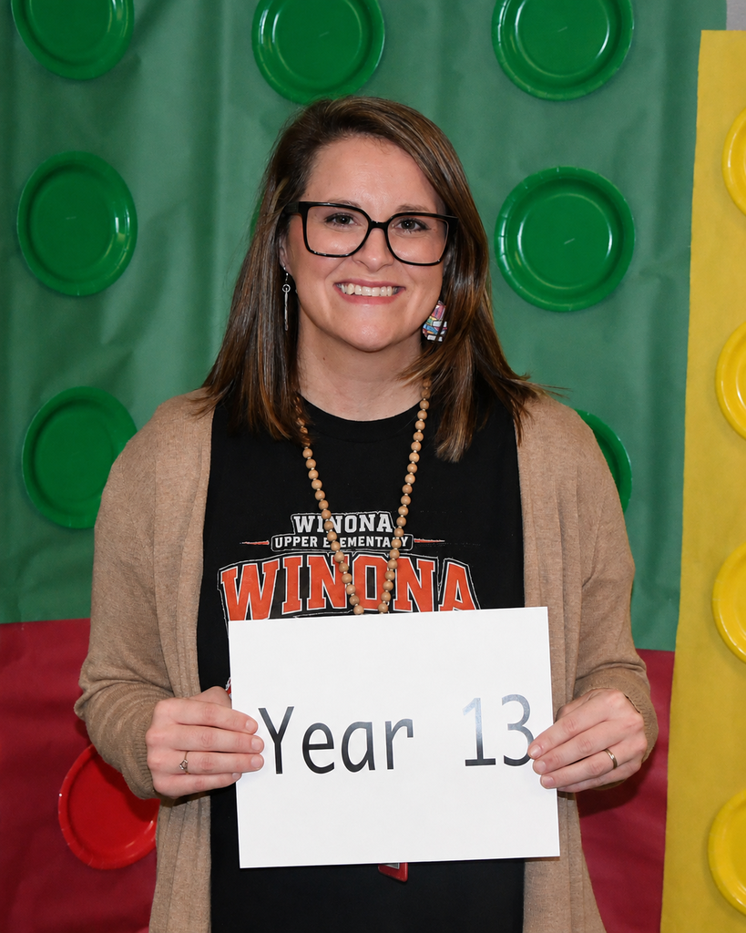 A woman with glasses and a necklace holds a sign that reads "Year 13" in front of a backdrop of green and yellow Lego pieces.