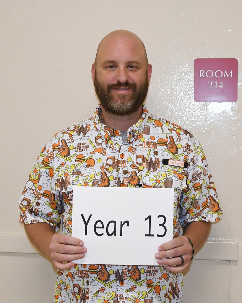 Man holding a white sign reading "Year 13" in a room with a door labeled "ROOM 214".