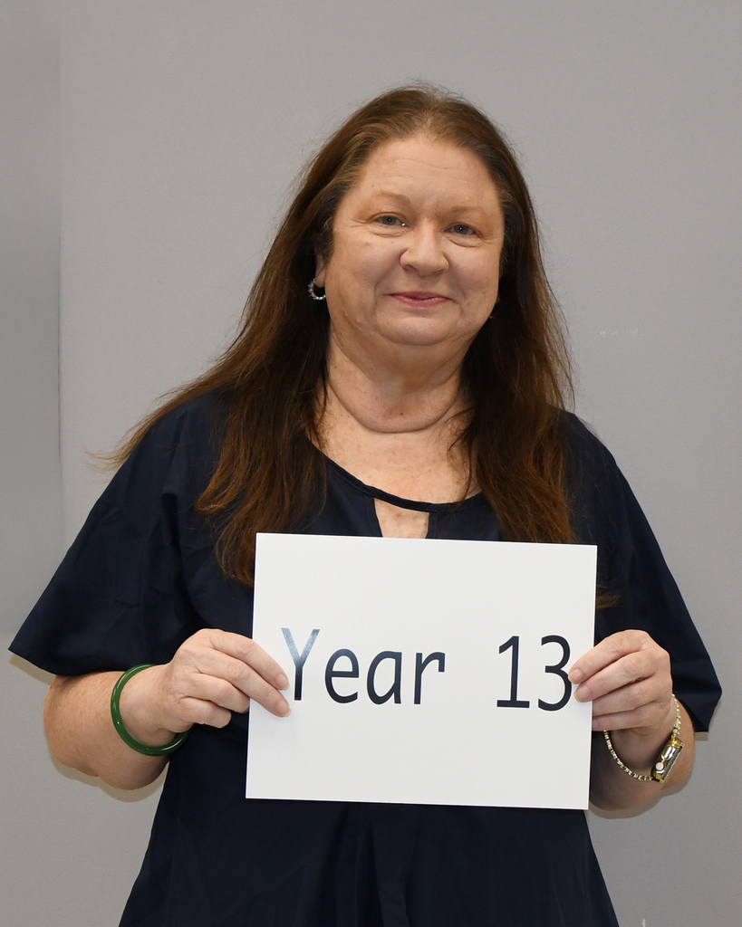 A woman with long hair, wearing a navy top, holds a sign reading "Year 13" against a gray background.