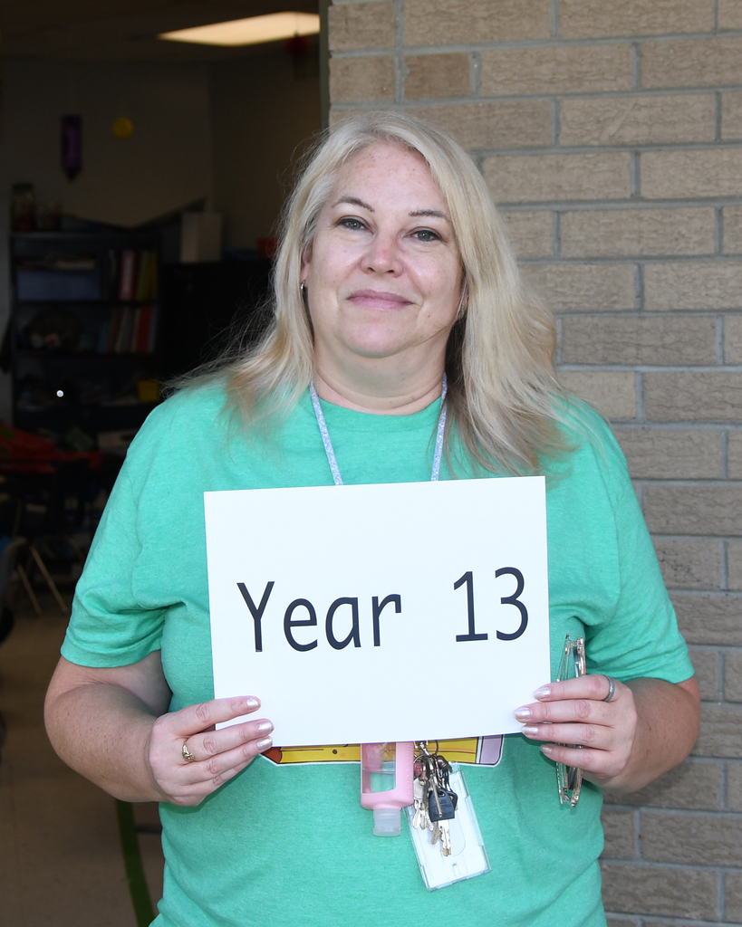 A woman in a green shirt holds a white sign that says "Year 13." She has blonde hair. Behind her is a brick wall.